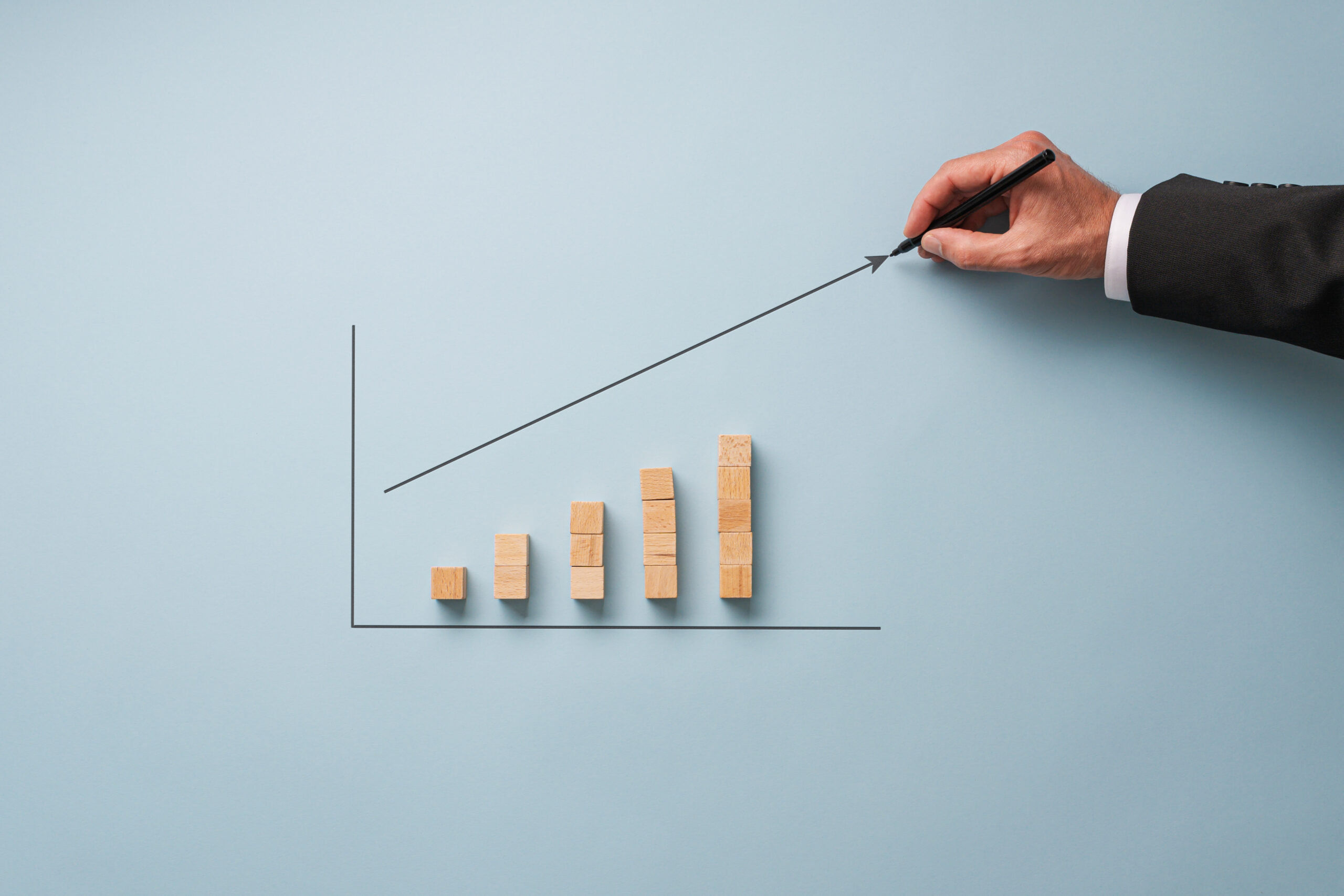 Hand of a businessman drawing a growing arrow above a column chart made of wooden blocks.
